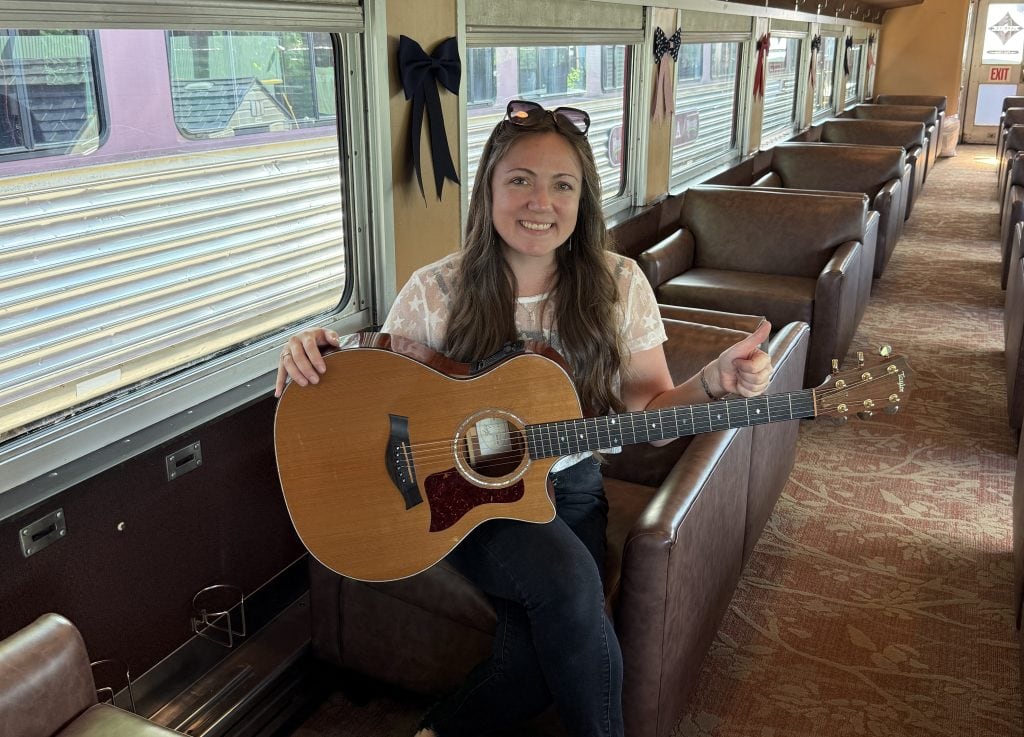 Woman with guitar inside vintage train car, smiling and giving a thumbs up.